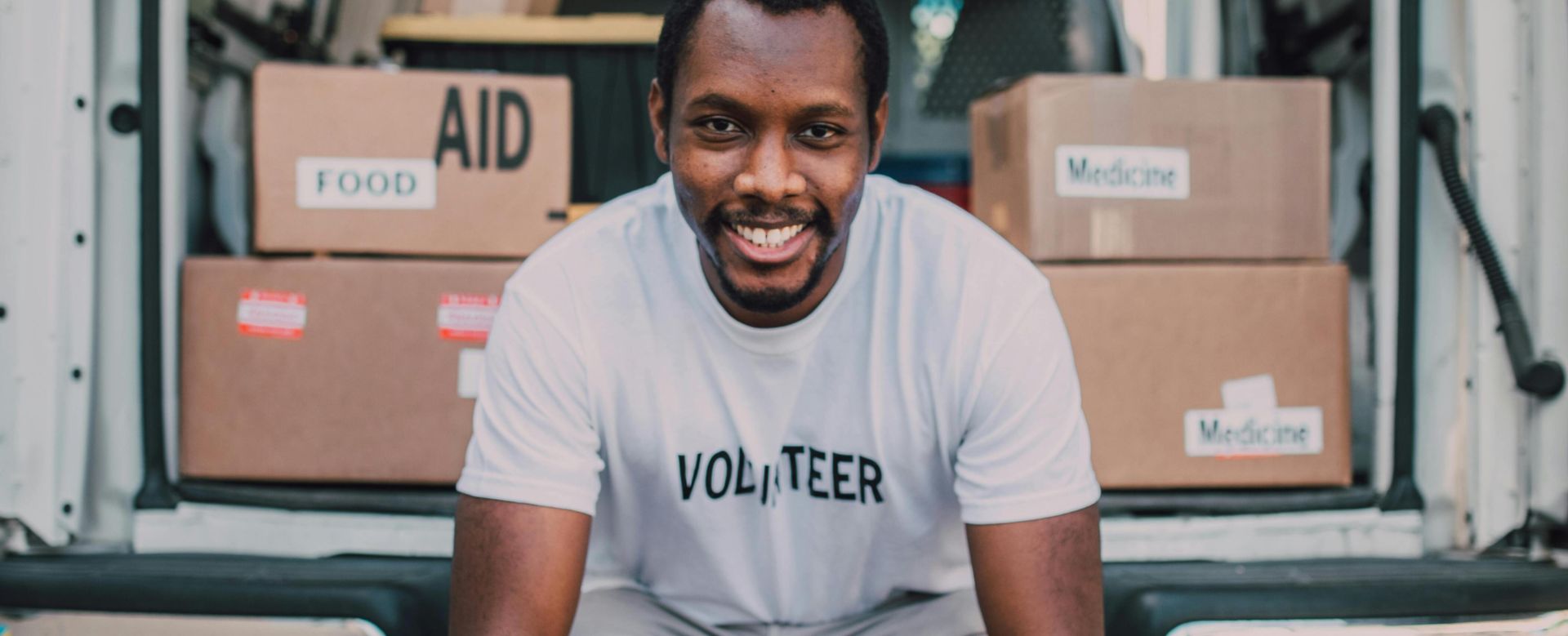 Smiling volunteer seated with boxes labeled food and medicine in a van.